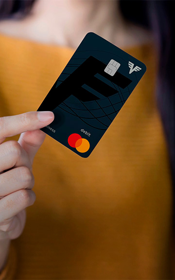 Eine Frau in einem gelben Pullover hält die Volksbank Debit Mastercard Business in der Hand. Man kann ihr Gesicht nicht sehen.