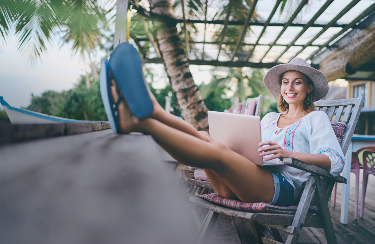 Eine Frau mit Sonnenhut sitzt in einem Sessel. Sie hat die Beine hochgelegt und ein Notebook auf dem Schoß.