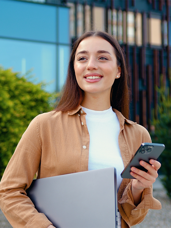 Eine junge Frau hat ein Notebook unter dem Arm und ein Mobiltelefon in der Hand. Sie lächelt.