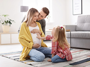 Eine schwangere Frau sitzt mit ihrem Mann und ihrer Tochter auf dem Boden. Das Mädchen berührt den Babybauch.