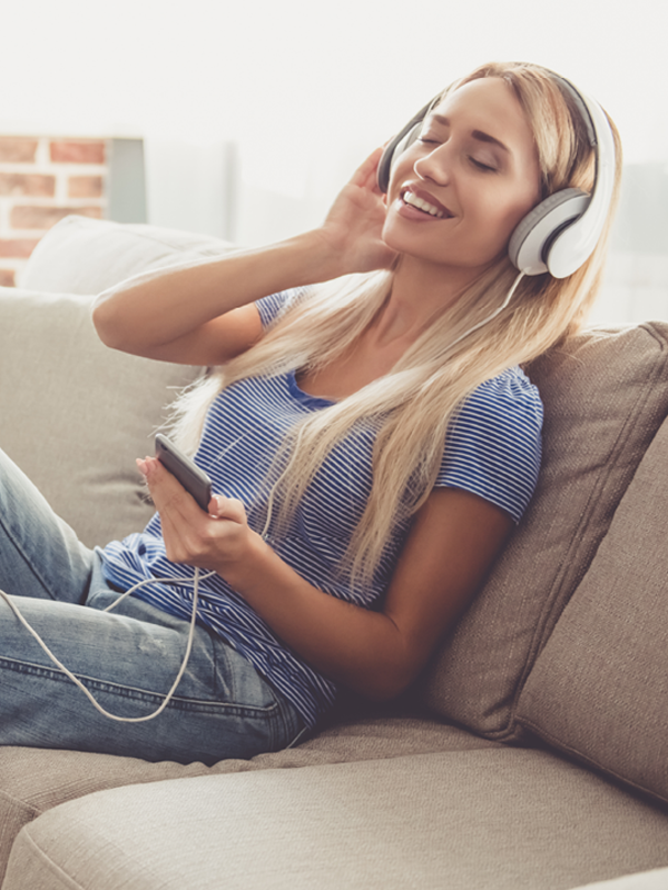 Eine Frau sitzt auf einer Couch und hört Musik über ihre Kopfhörer. Sie hat die Augen geschlossen und lächelt.