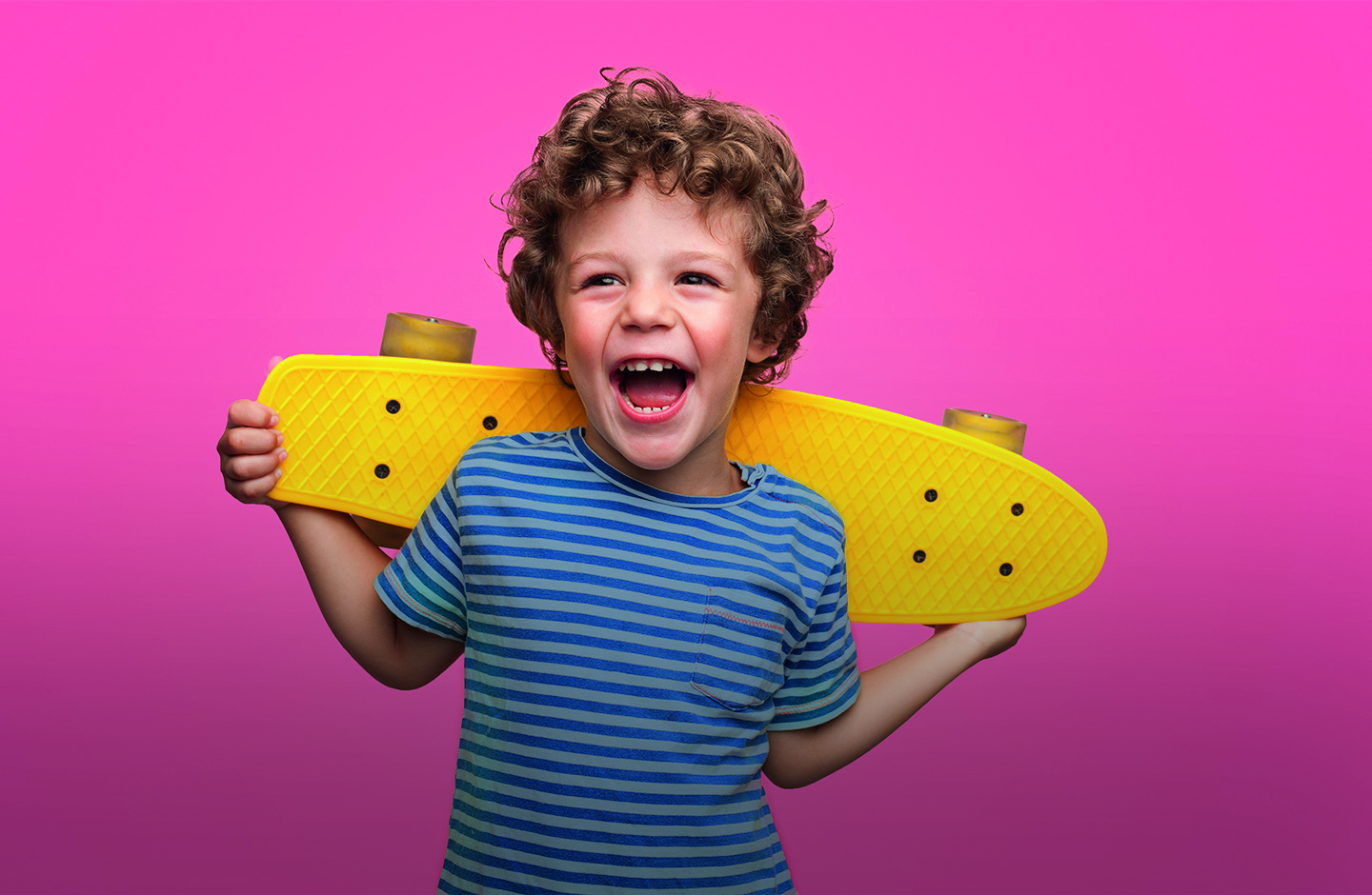 Ein Bub mit gelockten Haaren und einem gelben Skateboard lacht in die Kamera.
