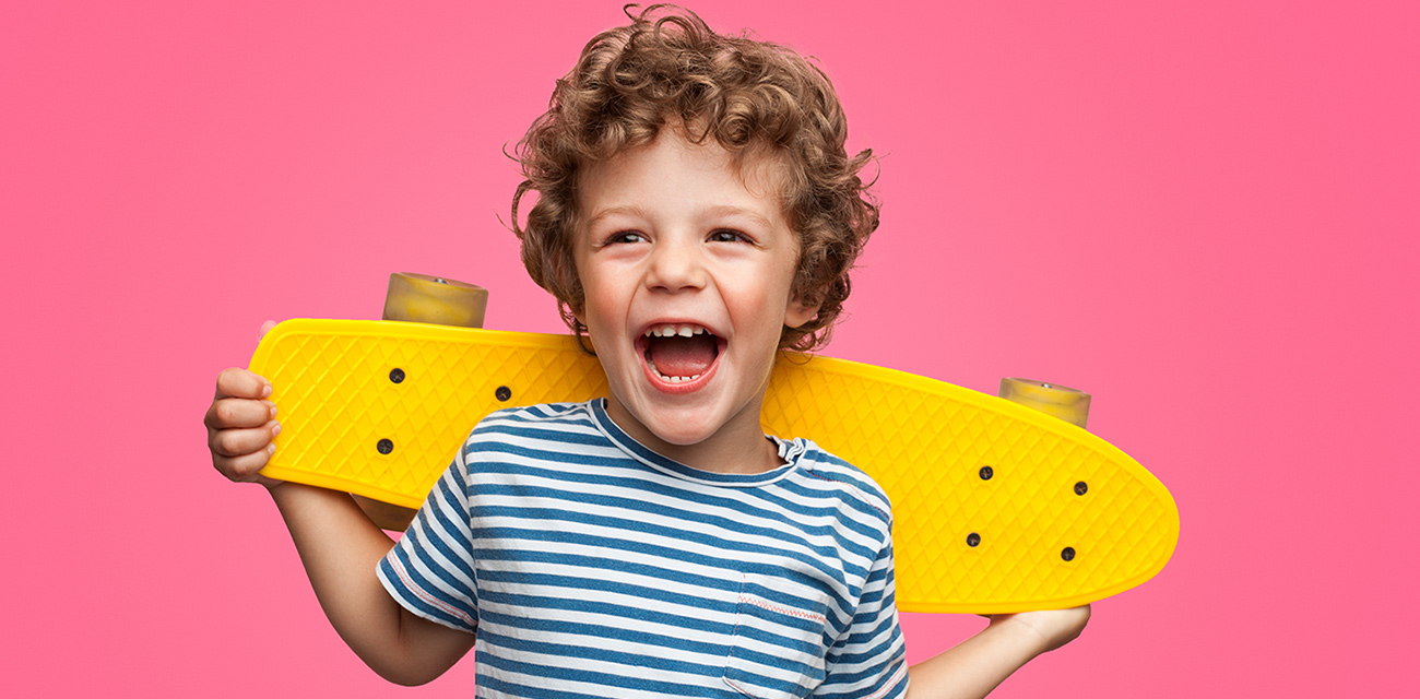 Ein kleiner Junge mit Locken lacht und hält ein gelbes Skateboard nach oben.