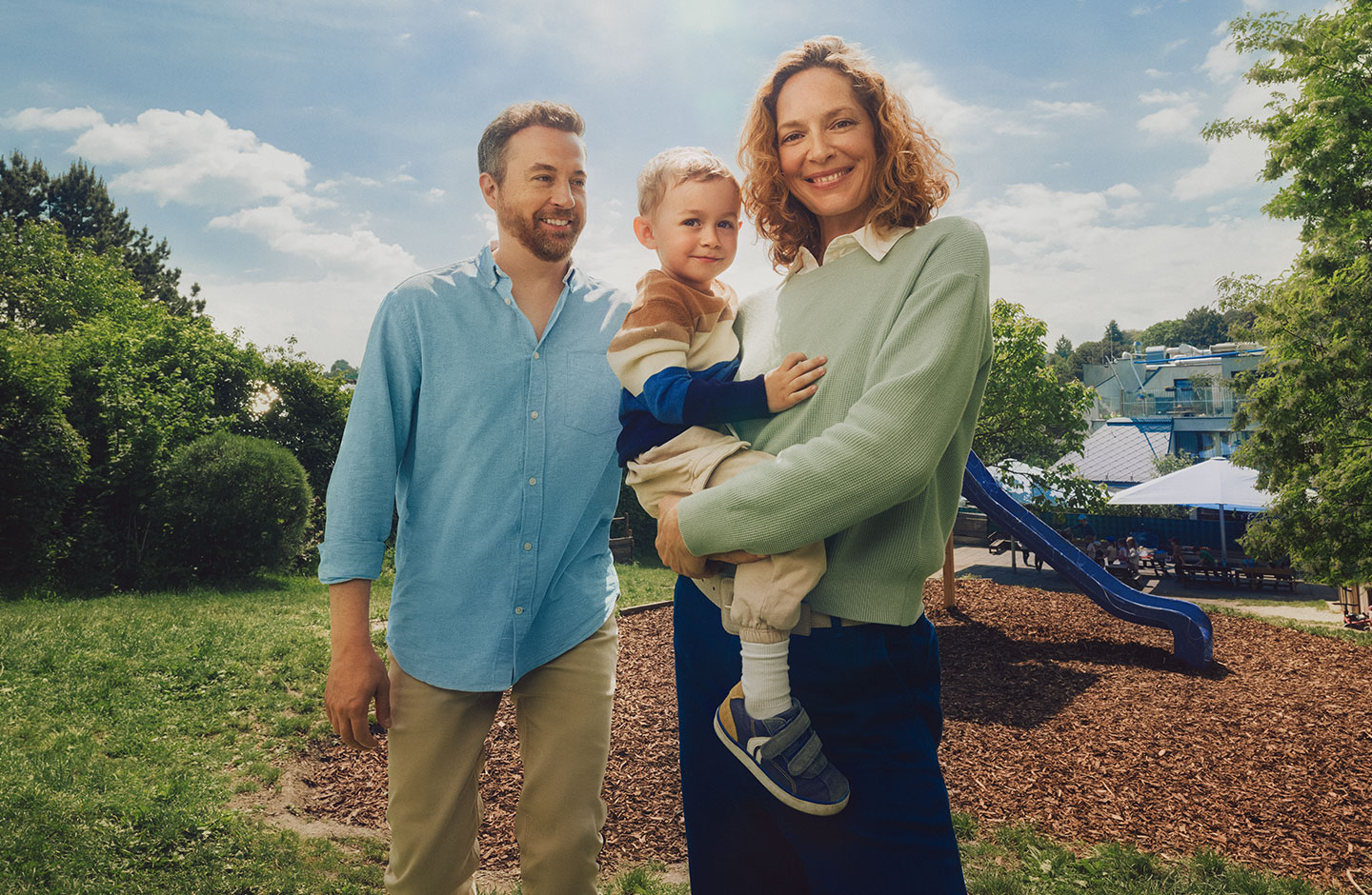 Eine jung Familie steht auf einem Spielplatz. Die Mutter hält das Kind auf dem Arm.
