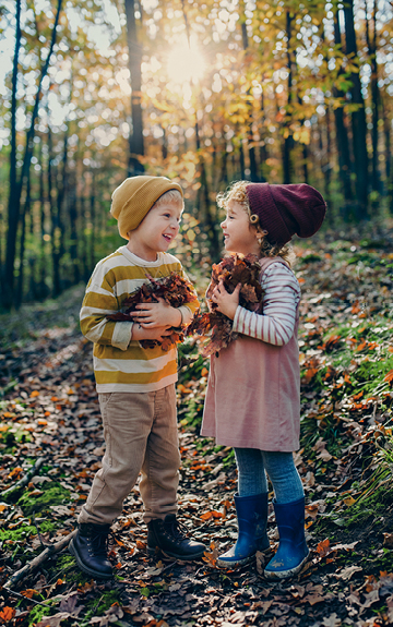 Zwei Kinder stehen in einem herbstlichen Wald. Beide haben trockene Blätter in den Armen.