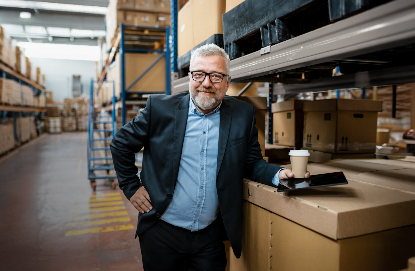 Ein Unternehmer steht in einer Lagerhalle. Er lehnt an einer Kiste und hat ein Tablet und einen Kaffeebecher aus Papier in der Hand.