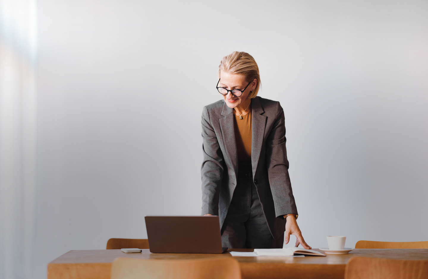Ältere Frau mit kurzen, blonden Haaren und Brille stützt sich auf einen Schreibtisch und blickt auf einen Laptop.