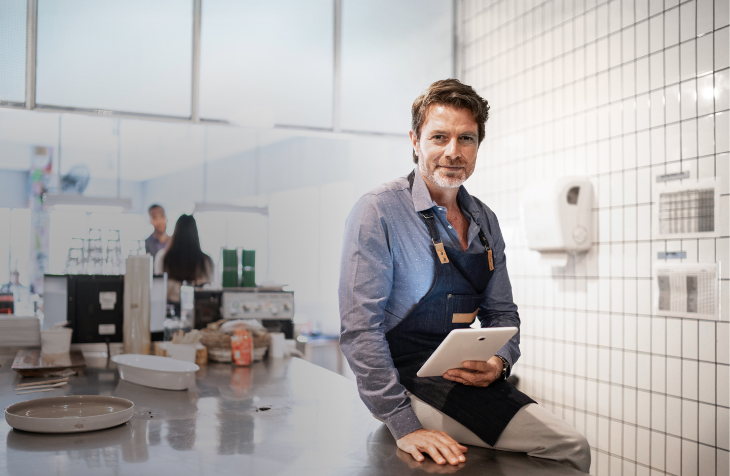 Ein Mann mit Schürze und Tablet in der Hand sitzt auf einer Arbeitsplatte in einer Küche und lacht.