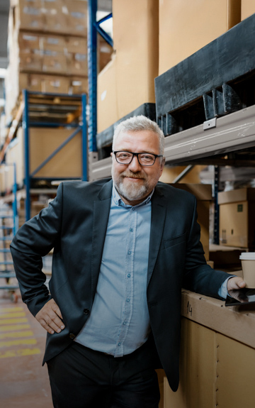 Ein Unternehmer steht in einer Lagerhalle. Er lehnt an einer Kiste und hat ein Tablet und einen Kaffeebecher aus Papier in der Hand.