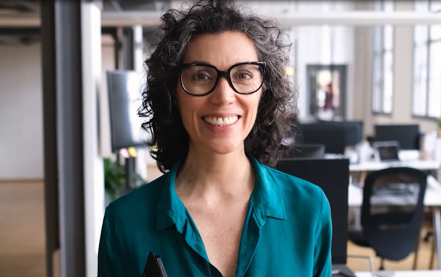Eine Frau mit lockigem Haar und Brille steht in einem Büro und lacht in die Kamera.