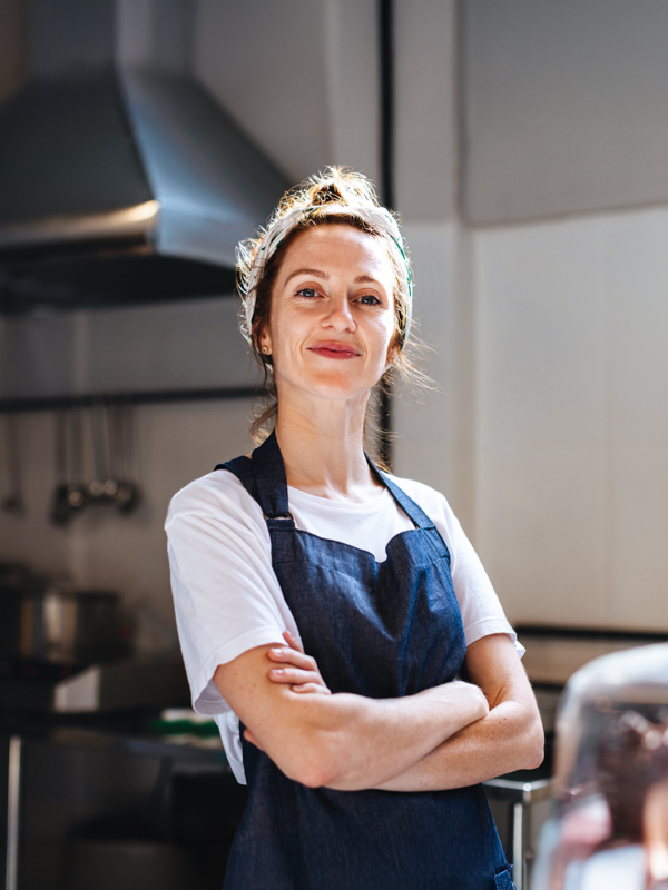 Eine junge Frau mit blondem Dutt und blauer Schürze verschränkt die Arme in einer Gastronomie-Küche und lächelt.