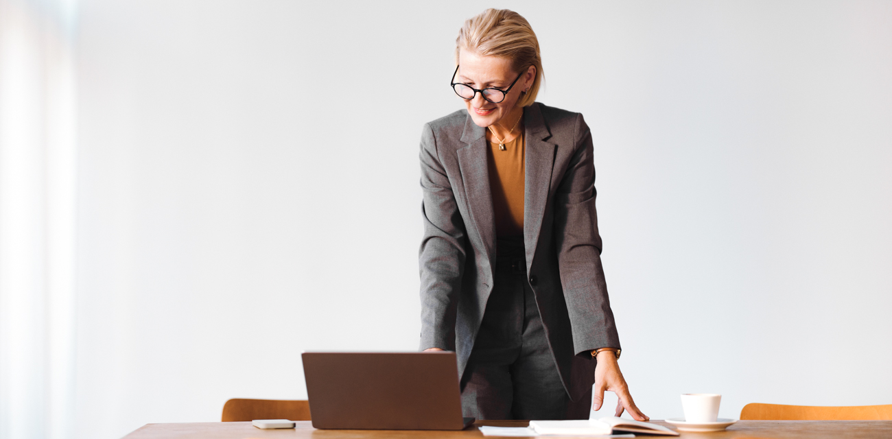 Eine ältere Dame mit kurzen blonden Haaren und Brille im Business-Anzug stützt sich mit den Händen auf einen Tisch und betrachtet lächelnd ihren Laptop.
