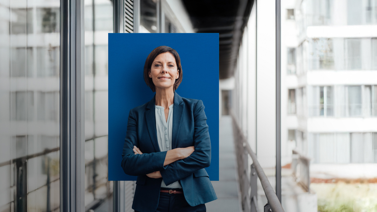 Eine Frau im Blazer verschränkt die Arme und steht auf einem gläsernen Büroflur. Hinter ihr befindet sich ein blaues Rechteck.