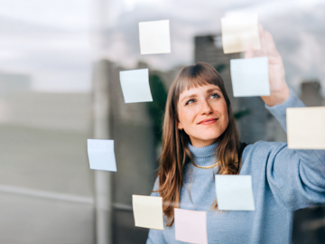 Eine Junge Frau mit blondem Haar klebt bunte Post-its an eine Fensterscheibe.