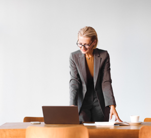 Ältere Frau stützt sich auf Schreibtisch und blickt lachend auf einen Laptop.