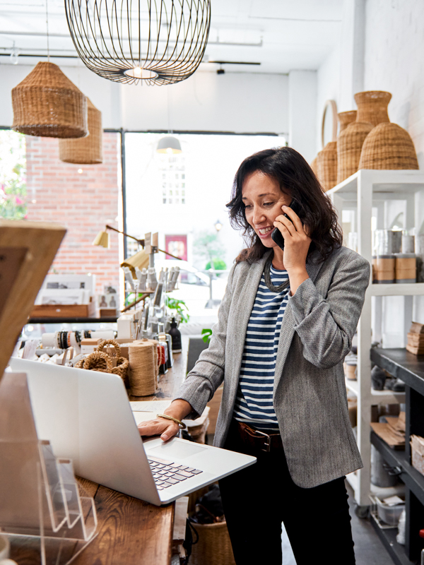 Eine Unternehmerin steht in ihrem Geschäft. Sie hat das Mobiltelefon am Ohr und blickt auf ein Notebook.