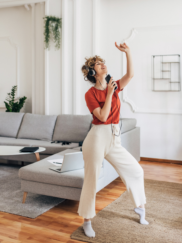 Eine Frau ist in ihrer Wohnung. Sie hat Kopfhörer auf und tantzt zur Musik von ihrem Mobiltelefon.