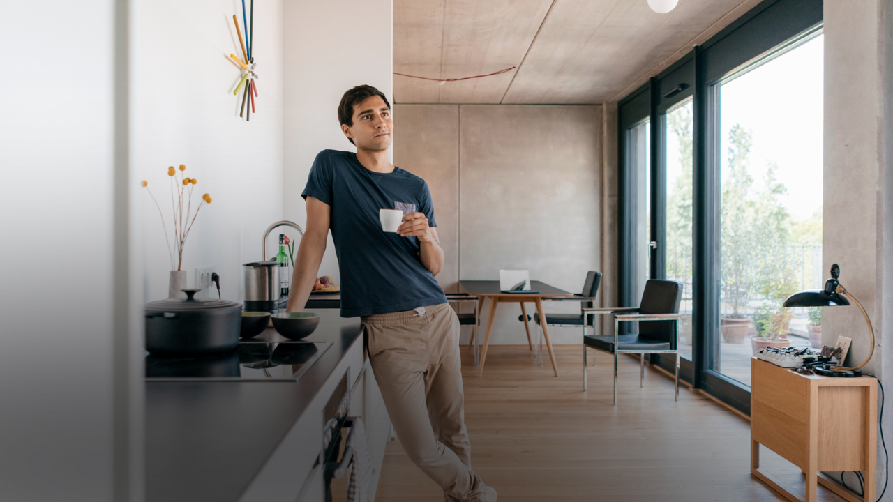 Ein Mann steht in einer Wohnküche. Er lehnt an der Küchenzeile und hält eine Kaffeetasse in der Hand.