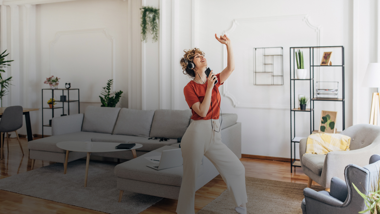 Eine Frau ist in ihrer Wohnung. Sie hat Kopfhörer auf und tantzt zur Musik von ihrem Mobiltelefon.