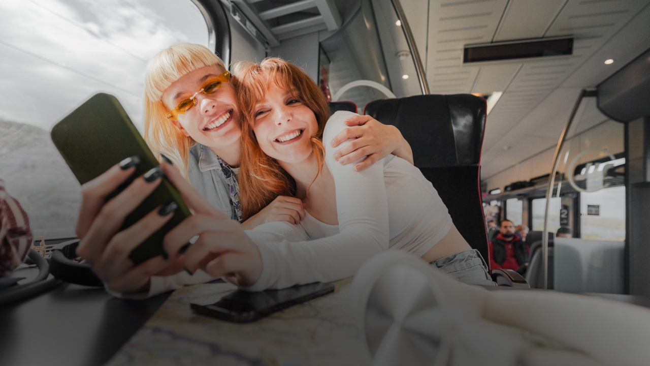 Zwei junge Frauen sitzen in einem Zug. Sie umarmen sich und blicken gemeinsam auf ein Mobiltelefon.