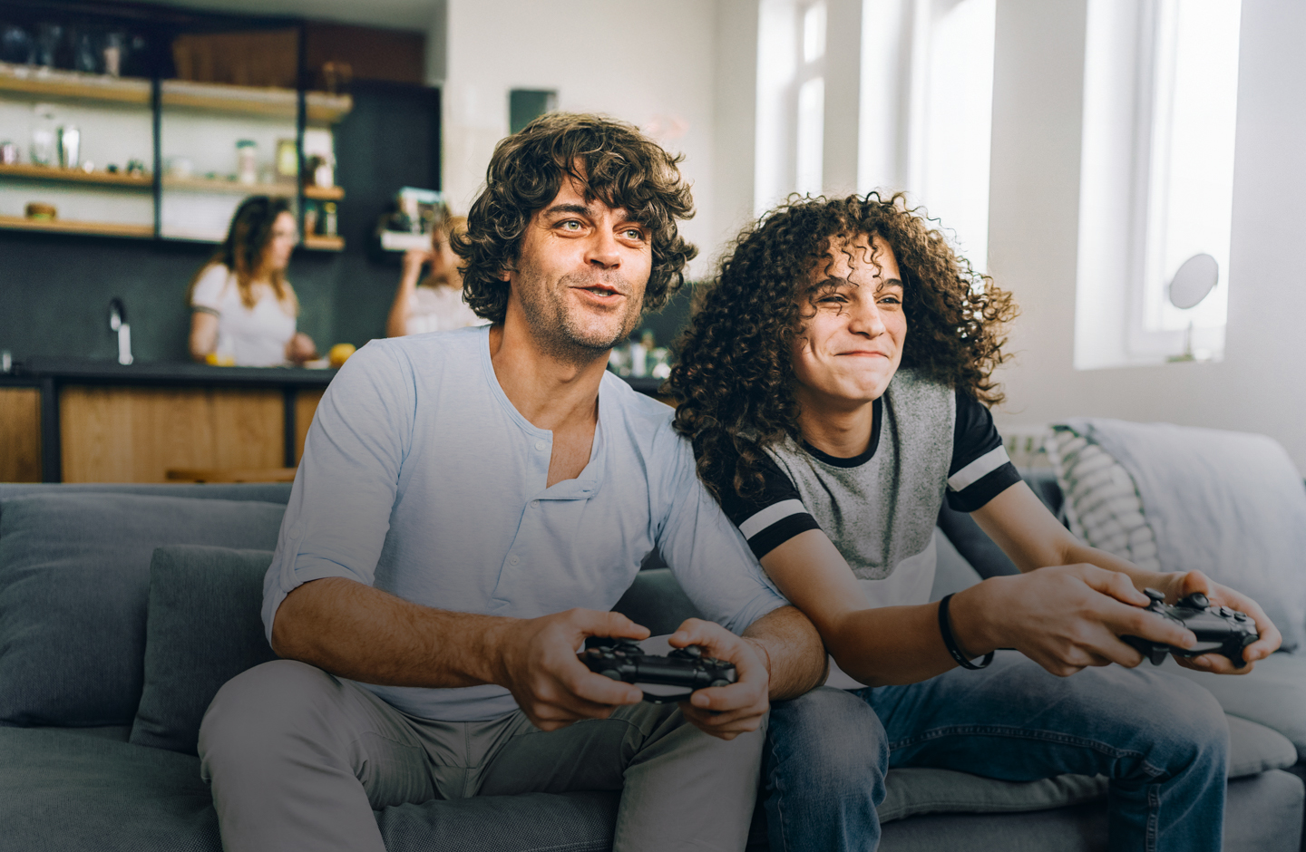 Vater und Sohn beim zocken mit Contollern in den Händen. Beide nah beieinander auf einem Sofa sitzend. Im Hintergrund verschwommen zwei Frauen in der Küche erkennbar.