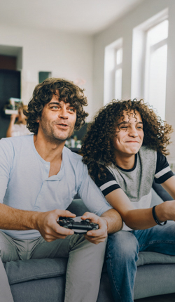 Ein Vater und sein Sohn sitzen auf einer Couch und spielen Videospiele mit Controller in der Hand.