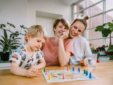 Eine Frau mit Kurzhaarfrisur sitzt mit ihren spielenden Kindern an einem Tisch.
