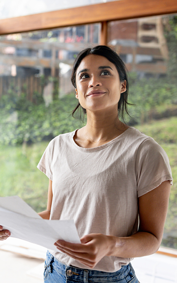 Eine junge Frau steht in ihrem neuen Haus, das gerade gebaut wird. Sie hat einen Bauplan in der Hand.