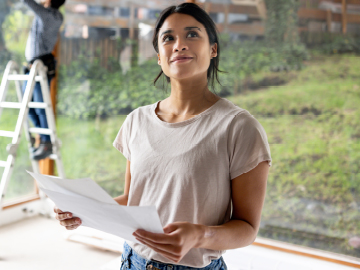 Eine junge Frau steht in ihrem neuen Haus, das gerade gebaut wird. Sie hat einen Bauplan in der Hand.