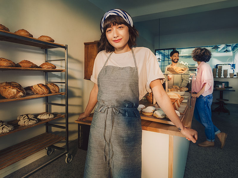 Eine Bäckerin steht in ihrer Backstube. Sie trägt eine graue Schürze und ein Kopftuch.