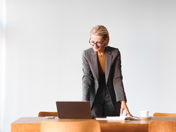 Eine ältere Frau mit Brille im Blazer stützt sich auf einen Tisch und blickt auf einen Laptop.